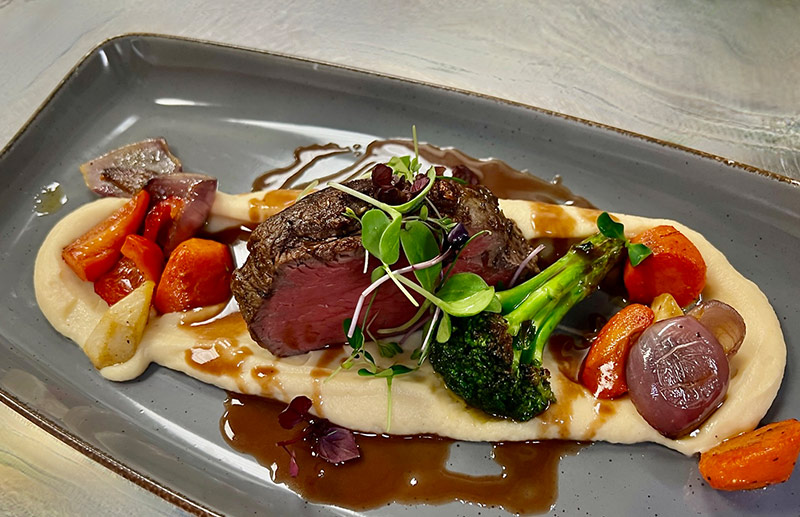 Zartes Rinderfilet auf cremigem Püree, mit Brokkoli, Karotten, roten Zwiebeln und Jus, serviert auf grauer Porzellanplatte.