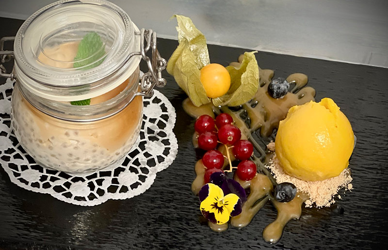Hausgemachtes Dessert im Glas mit Minze, dazu Beeren, Physalis, essbare Blüten und Sorbet, kunstvoll angerichtet auf Schieferplatte.