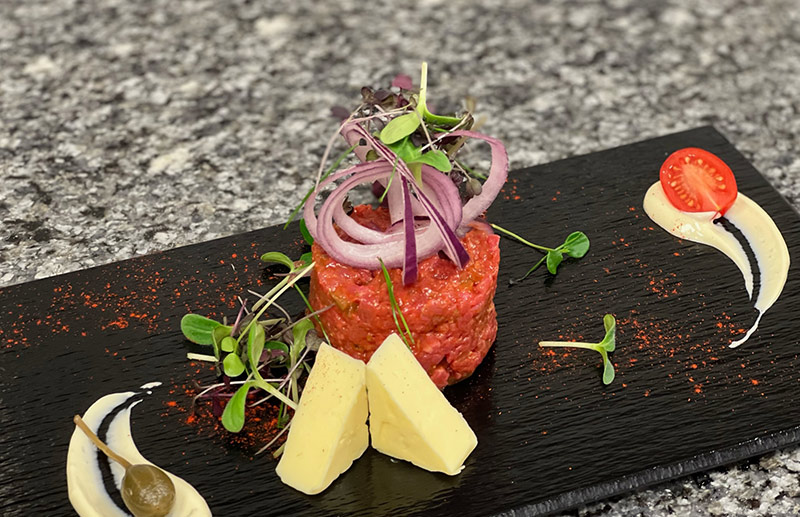 Rindertatar auf Schieferplatte, garniert mit roten Zwiebeln, Microgreens und Käsestücken.